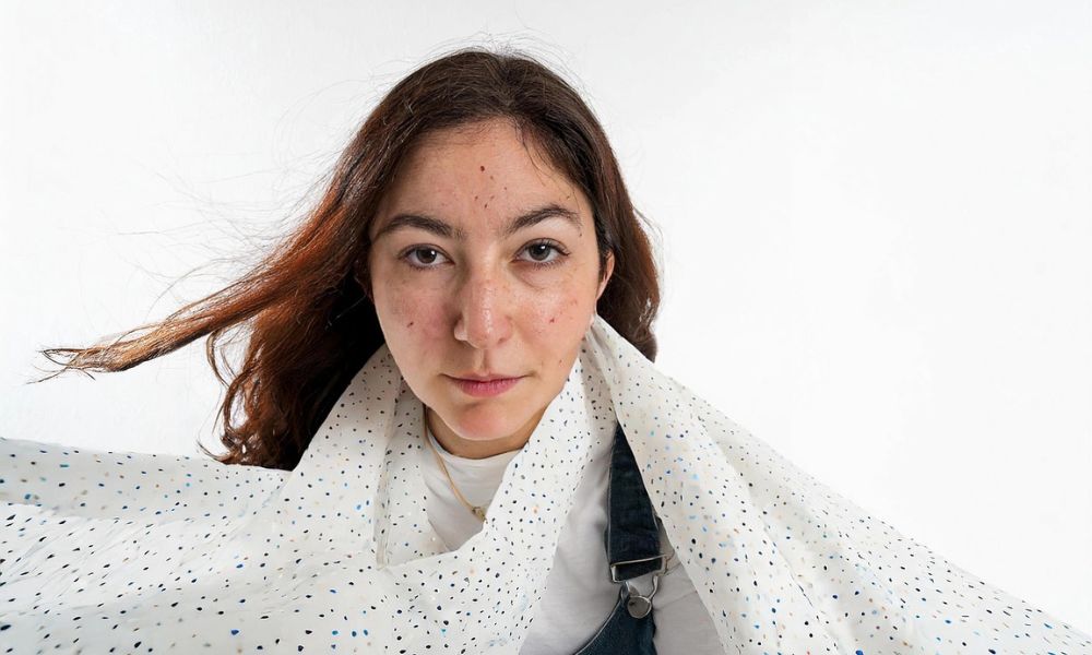 Une personne aux cheveux longs regarde l'objectif, drapée dans un tissu léger à pois sur un fond uni. Sa peau rayonne naturellement, révélant peut-être le secret d'un sérum anti-taches pour un look impeccable.