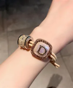 Gros plan d'un poignet portant un bracelet en or avec une breloque carrée comportant la lettre « D » et d'autres éléments décoratifs ronds.