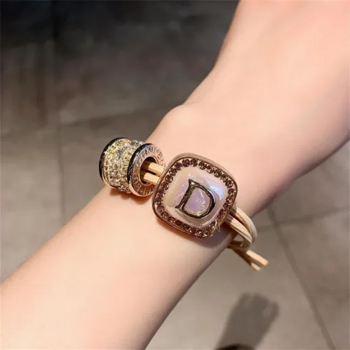 Gros plan d'un poignet portant un bracelet en or avec une breloque carrée comportant la lettre « D » et d'autres éléments décoratifs ronds.