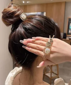 Une personne coiffée en chignon tient sa main près de sa tête, mettant en valeur deux bagues et de longs ongles décorés. L'arrière-plan semble être un espace intérieur avec des éléments en bois.