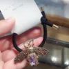 Une main tient l'une des Épingles à Cheveux Rétro Élégantes, un élastique noir orné d'un pendentif abeille en bronze orné d'une pierre violette. L'élastique, qui rappelle le charme rétro, est attaché à un emballage sur lequel est inscrit « Fabriqué en Corée ».