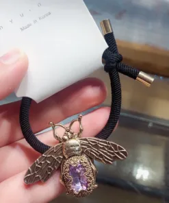 Une main tient l'une des Épingles à Cheveux Rétro Élégantes, un élastique noir orné d'un pendentif abeille en bronze orné d'une pierre violette. L'élastique, qui rappelle le charme rétro, est attaché à un emballage sur lequel est inscrit « Fabriqué en Corée ».