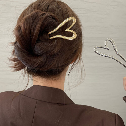 Vue de dos d'une personne aux cheveux châtains coiffés en chignon, maintenus par une pince à cheveux en métal en forme de cœur, portant une veste marron. Ils tiennent un accessoire pour cheveux similaire en forme de cœur dans une main.