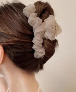 Gros plan de l'arrière de la tête d'une femme aux cheveux bruns coiffés en chignon élégant, ornés d'un chouchou à volants de couleur claire. La personne porte également des boucles d'oreilles en perles et a la Grande Épingle à Cheveux en Maille pour Femmes délicatement glissée dans le chignon.