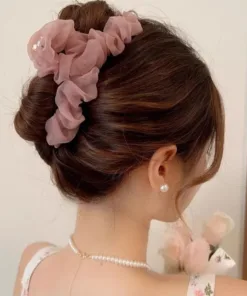 Une femme aux cheveux châtains coiffés en chignon élégant porte un accessoire de coiffure à volants rose et des boucles d'oreilles en perles, avec quelques fleurs visibles en arrière-plan. Son choix de Grande Épingle à Cheveux en Maille pour Femmes ajoute une touche de sophistication à son look.