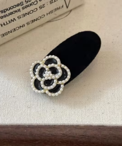 Une Petite pince à cheveux élégante en perles en noir, comportant une fleur ornée de perles, placée sur une surface en tissu de couleur claire près d'une boîte partiellement visible.