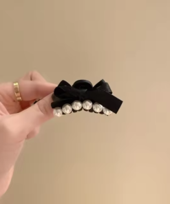 Une main tient la Petite pince à cheveux de mode coréenne, une pince à cheveux chic en forme de nœud noir ornée d'une rangée de perles blanches, mettant en valeur un accessoire élégant.