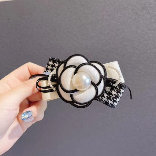 Une main aux ongles peints tient une Pince à Cheveux Fleur de Camélia Classique ornée d'un centre en fausse perle et d'un ruban à motif pied-de-poule.