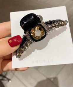 Une main avec du vernis à ongles rouge tient un « Pinces à Cheveux Rétro Élégantes – Épingles en Clip avec Strass pour Femmes et Filles » noir, orné d'une grande pierre précieuse centrale et d'une rangée de strass plus petits, positionnés sur une carte d'affichage blanche avec un texte pâle .