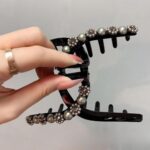 Une main avec une bague en or tient une pince à cheveux noire en forme de crabe, ornée de perles artificielles et de strass, sur un fond uni.