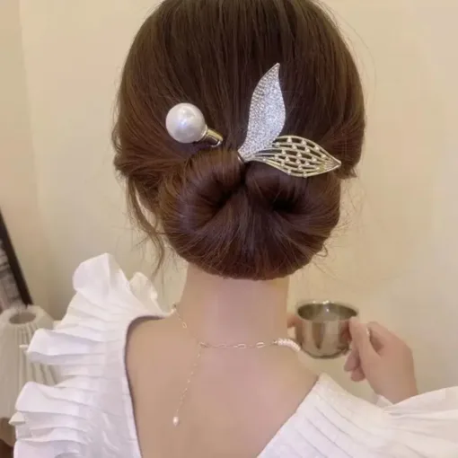 Une femme avec un chignon soigneusement coiffé à l'aide du Rouleau de bandeau en forme de beignet pour chignon, orné d'un accessoire pour cheveux en perles et feuilles, portant un haut blanc à manches à volants, tient une petite tasse dans sa main droite.