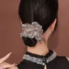 Une femme aux cheveux noirs coiffés en chignon bas, ornée d'un grand chouchou décoratif, tourne le dos à la caméra. Elle porte un haut noir décoré de détails argentés autour de l'encolure. Cette image ressemble presque à un AUTO-DRAFT d'élégance et de simplicité.