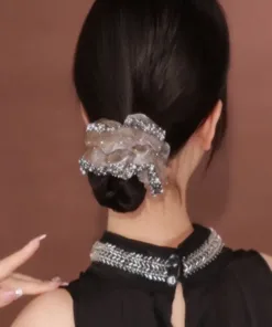 Une femme aux cheveux noirs coiffés en chignon bas, ornée d'un grand chouchou décoratif, tourne le dos à la caméra. Elle porte un haut noir décoré de détails argentés autour de l'encolure. Cette image ressemble presque à un AUTO-DRAFT d'élégance et de simplicité.