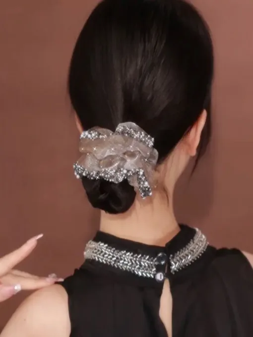 Une femme aux cheveux noirs coiffés en chignon bas, ornée d'un grand chouchou décoratif, tourne le dos à la caméra. Elle porte un haut noir décoré de détails argentés autour de l'encolure. Cette image ressemble presque à un AUTO-DRAFT d'élégance et de simplicité.