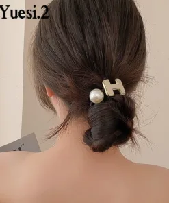 Une personne aux cheveux bruns coiffés en chignon bas, maintenus par une épingle à cheveux AUTO-DRAFT ornée d'un « H » doré et d'un ornement en perles. Le fond est uni.