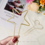 Une main tenant un Bâton à cheveux en métal en forme d'étoile simple. En arrière-plan, il y a un marque-page en métal en forme de cœur, un livre et une composition florale.