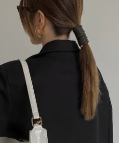 Une femme aux cheveux bruns attachés en queue de cheval basse, portant des lunettes de soleil et un blazer noir, porte un sac à main blanc sur son épaule, dégageant un style chic AUTO-DRAFT.