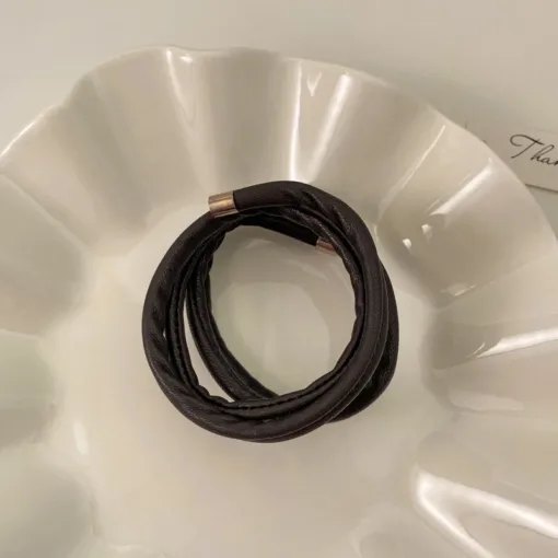 Un bracelet en cuir noir spiralé Chouchous en cuir INS Temperament avec un fermoir en métal repose sur une plaque blanche décorative, ajoutant un élément d'élégance à la monture.