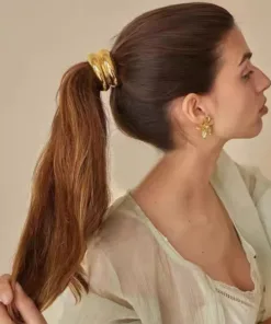 Une personne aux cheveux bruns attachés en queue de cheval par des anneaux dorés, portant un haut clair et des boucles d'oreilles dorées en forme de fleur, regarde vers la droite sur un fond uni AUTO-DRAFT.