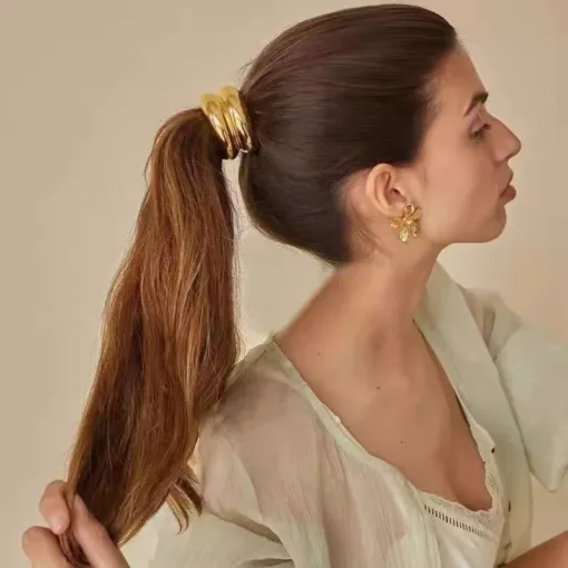 Une personne aux cheveux bruns attachés en queue de cheval par des anneaux dorés, portant un haut clair et des boucles d'oreilles dorées en forme de fleur, regarde vers la droite sur un fond uni AUTO-DRAFT.