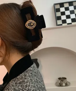 Une personne aux cheveux châtains coiffés en chignon, maintenus avec la Pince à Cheveux en Velours Flocage Rose Noir, ornée d'un emblème doré, est partiellement visible. À l'arrière-plan, on distingue un cadre à carreaux noir et blanc et une petite tasse métallique.