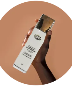 Une main tient gracieusement un flacon de lotion pour le corps Nouveau Belle Nubian Caramel, orné d'un bouchon doré, sur fond beige apaisant.