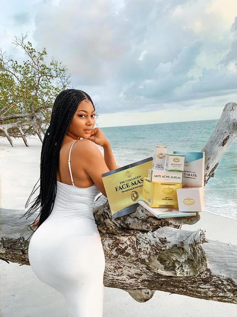 Une femme en robe blanche pose devant un arbre sur une plage, présentant des produits de soins pour la peau. L'océan et le ciel nuageux sont en arrière-plan.