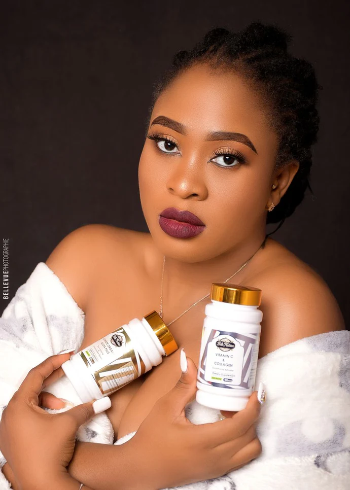 Une femme en robe blanche tient avec assurance deux flacons étiquetés « PILULES BIO ULTIMES + VITAMINE C » et « COLLAGÈNE (le set le plus éclairant) » avec des couvercles dorés, posant sur un fond sombre. Une femme en robe blanche tient avec assurance deux flacons étiquetés "PILULES BIO ULTIMES + VITAMINE C" et "COLLAGÈNE (le set le plus éclairant)" avec des couvercles dorés, posant sur un fond sombre.