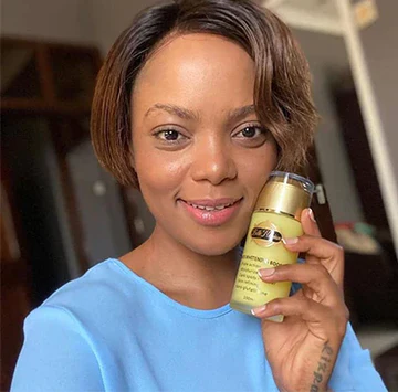 Une femme aux cheveux courts portant un haut bleu clair brandit un flacon jaune de produit de soin et sourit à la caméra à l'intérieur.