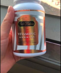 Hand holding a white bottle of Vitamin C Gummies with an orange label near a window.