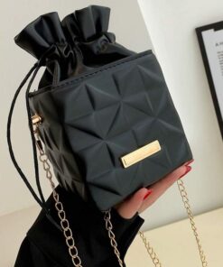 Black quilted drawstring handbag with a gold chain strap, held by a person with red nails; gold plaque on front.