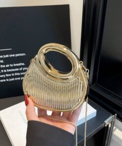 Gold metallic circular handbag with a ribbed body and large round handle, held by a hand with red nails and a thin gold chain strap visible.